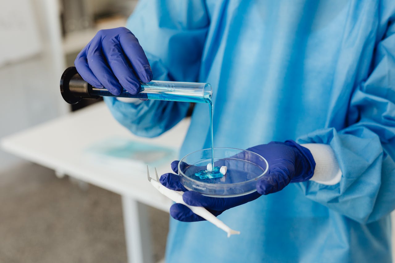 Scientist in lab gown pouring blue liquid from test tube into petri dish wearing gloves.